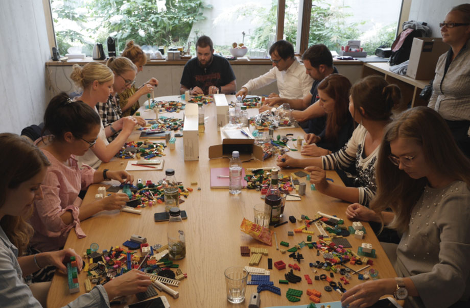 12 Personen sitzen um einen Tisch und bauen mit Legosteinen