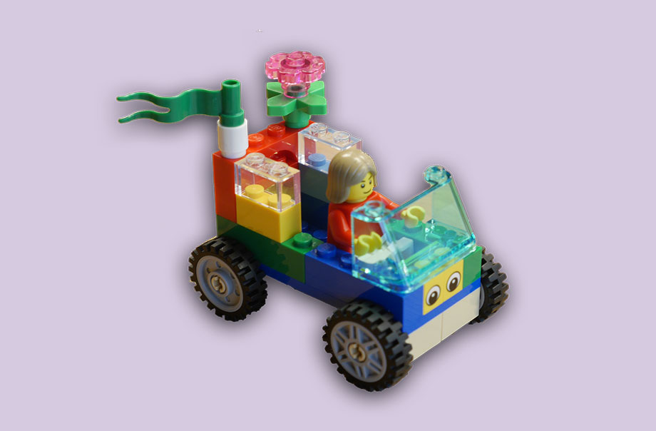 Selbstgebautes Lego Auto aus bunten Steinen mit einer Fahne, Blume und ohne Dach. Fahrzeuglenker mit langen braunen Haaren.